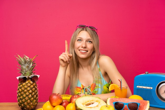 Joven rubia con muchas frutas señalando con el dedo índice una gran idea