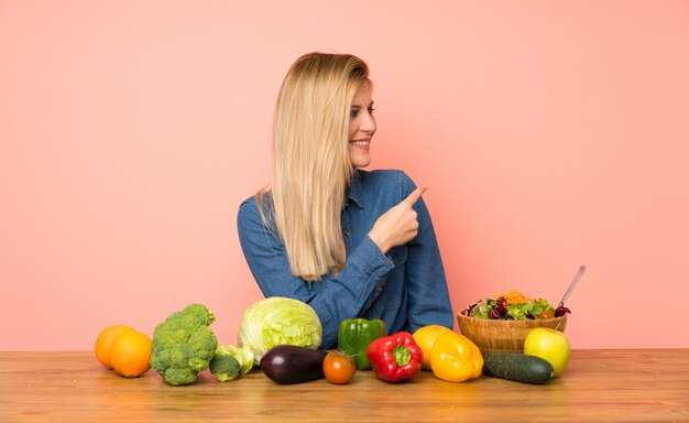 Joven rubia con muchas verduras apuntando hacia un lado para presentar un producto