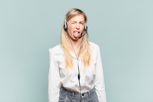 Joven rubia mujer de telemercadeo que se siente disgustada e irritada, sacando la lengua, disgustando algo desagradable y asqueroso