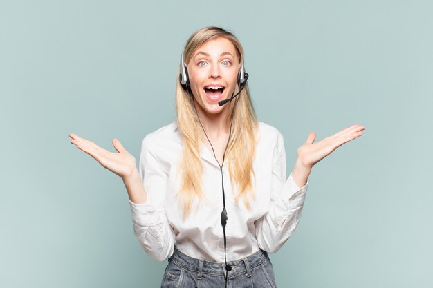 Joven rubia mujer de telemercadeo que se siente feliz, emocionada, sorprendida o conmocionada, sonriendo y asombrada por algo increíble