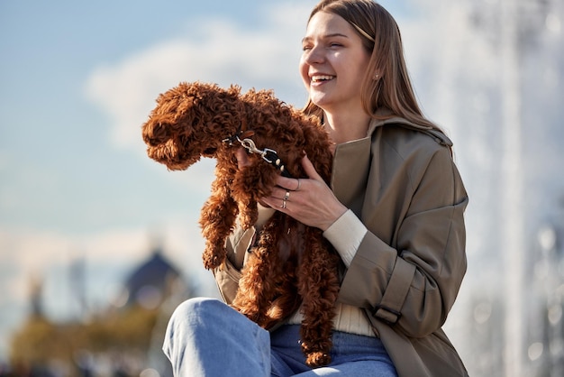 Una joven rubia muy sonriente con un impermeable beige se sienta en un banco en el parque y sostiene su corte