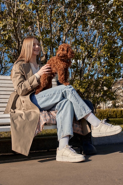 Una joven rubia muy sonriente con un impermeable beige se sienta en un banco en el parque y sostiene su corte