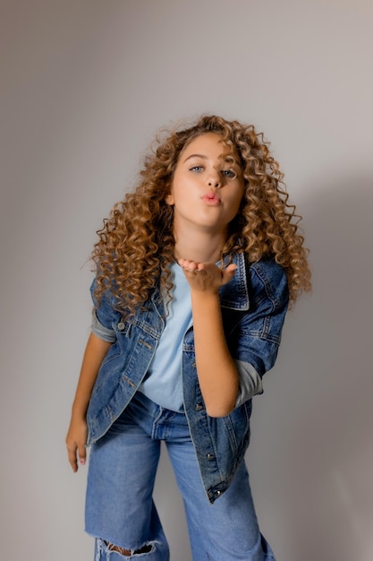 Joven rubia con ojos azules y cabello rizado en una chaqueta de mezclilla y jeans envía un beso al aire