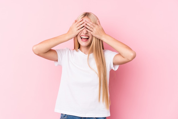 Joven rubia en pared rosa cubre los ojos con las manos, sonríe ampliamente esperando una sorpresa.