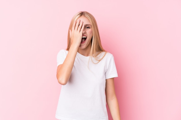 Joven rubia en pared rosa divirtiéndose cubriendo la mitad de la cara con la palma.