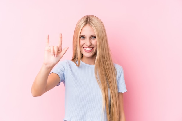 Joven rubia en pared rosa mostrando un gesto de cuernos