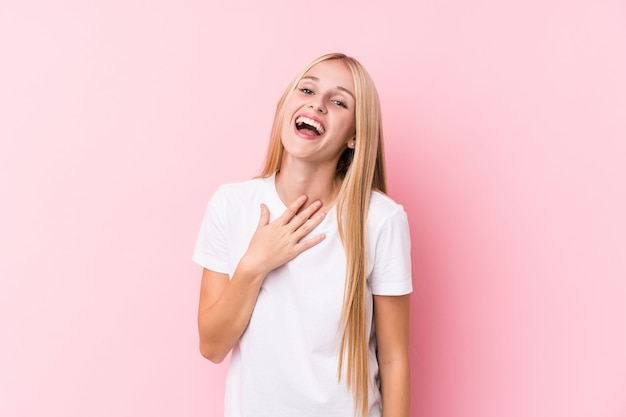 Joven rubia en pared rosa se ríe a carcajadas manteniendo la mano en el pecho.