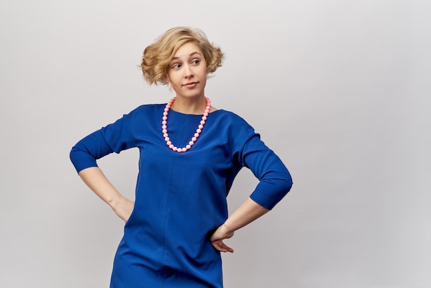 Joven rubia con pelo corto y rizos, posando para el estudio de rodaje. Lleva un vestido azul y cuentas vintage. Expresión facial amigable y mirada a un lado. Las manos descansaban sobre sus caderas.