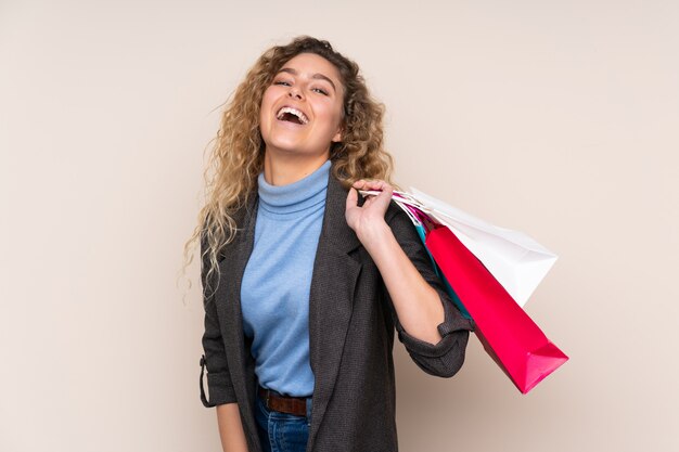 Joven rubia con el pelo rizado aislado en la pared de color beige con bolsas de compras y sonriendo