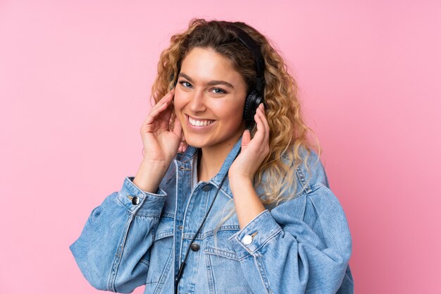 Joven rubia con el pelo rizado aislado en la pared rosa escuchando música