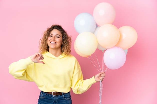 Joven rubia con el pelo rizado atrapando muchos globos en la pared rosa orgullosa y satisfecha