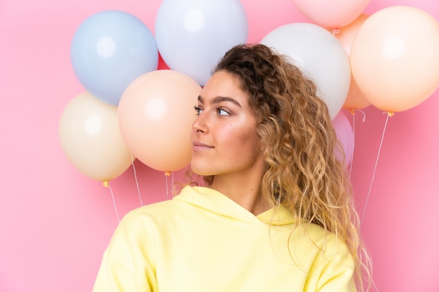 Joven rubia con el pelo rizado atrapar muchos globos en la pared rosa