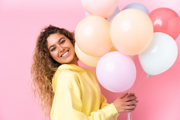 Joven rubia con el pelo rizado atrapar muchos globos en la pared rosa