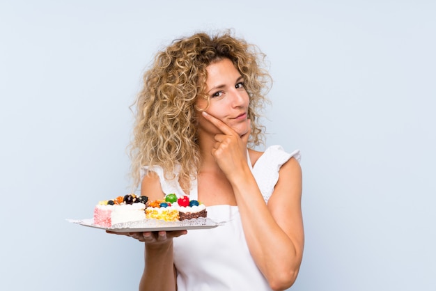 Joven rubia con el pelo rizado con muchos mini pasteles diferentes pensando en una idea