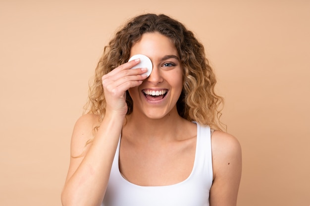 Foto joven rubia con el pelo rizado en la pared de color beige con algodón para quitar el maquillaje de la cara y sonriendo