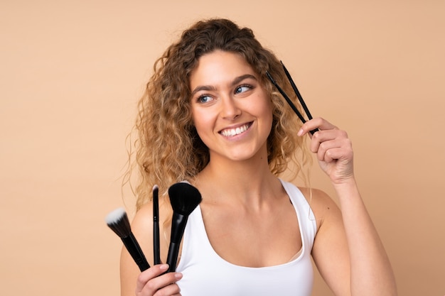 Joven rubia con el pelo rizado en la pared de color beige con pincel de maquillaje