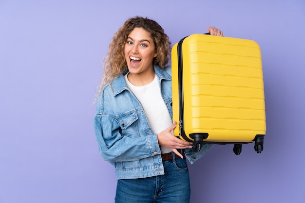 Foto joven rubia con el pelo rizado en la pared de color púrpura en vacaciones con maleta de viaje