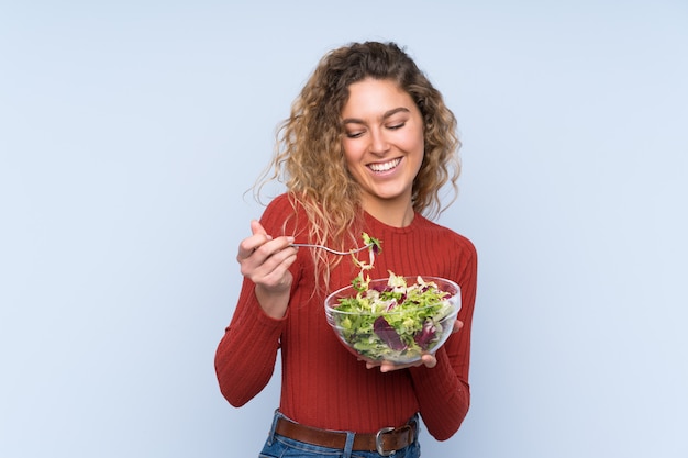 Foto joven rubia con el pelo rizado sosteniendo una ensalada sobre pared aislada