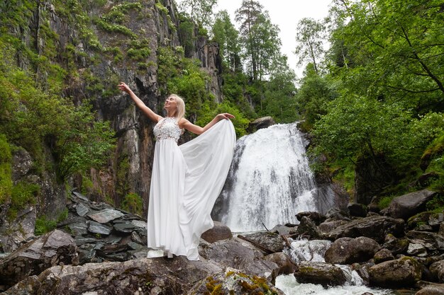 Foto una joven rubia en una pose elegante levanta un vestido boudoir en las montañas contra una cascada y piedras levantando las manos como una bailarina en un cálido día de verano en altai con árboles verdes