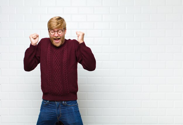 Joven rubia que parece extremadamente feliz y sorprendida, celebrando el éxito, gritando y saltando contra la pared de azulejos vintage