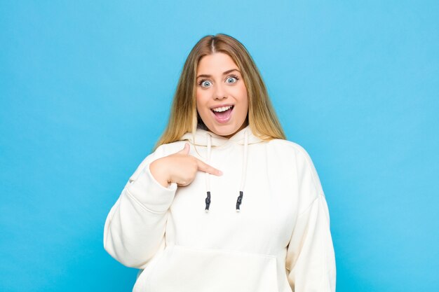 Joven rubia que parece feliz, orgullosa y sorprendida, alegremente apuntando a sí misma, sintiéndose segura y elevada contra la pared plana