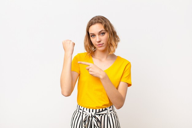 Joven rubia que parece impaciente y enojada, apuntando al reloj, pidiendo puntualidad, quiere llegar a tiempo contra la pared de color plano