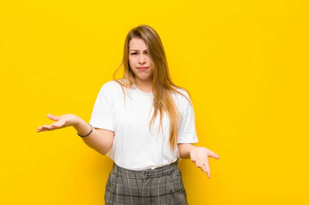 Joven rubia que parece perpleja, confundida y estresada, preguntándose entre diferentes opciones, sintiéndose insegura contra la pared naranja