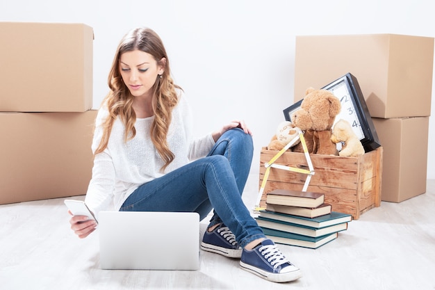 Una joven rubia renovando su casa sentada en el suelo rodeada de cajas de cartón con un tablet-pc