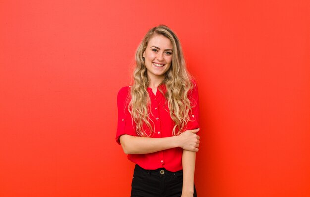 Joven rubia riendo con timidez y alegría, con una actitud amistosa y positiva pero insegura en la pared roja