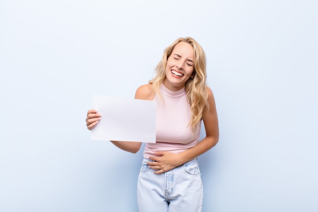 Joven rubia riéndose a carcajadas de alguna broma hilarante, sintiéndose feliz y alegre, divirtiéndose sosteniendo una hoja de papel