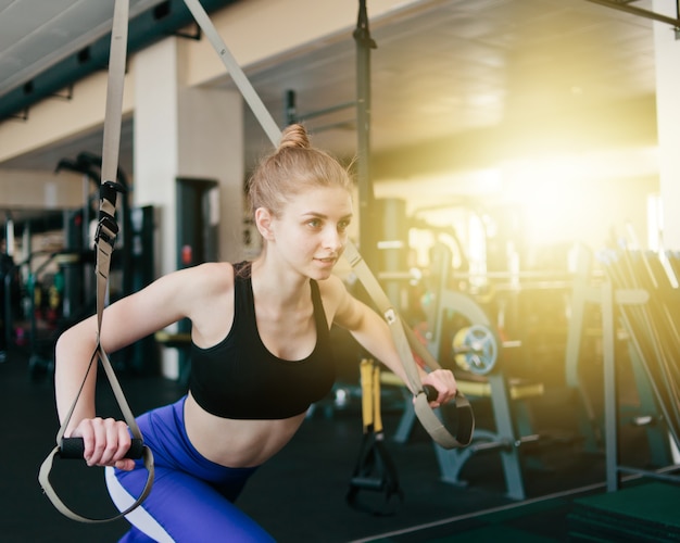 Joven rubia en ropa deportiva haciendo ejercicio con correas de fitness en el gimnasio. Entrenamiento funcional