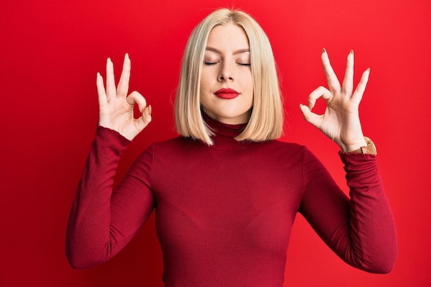 Joven rubia con ropa informal relajada y sonriendo con los ojos cerrados haciendo gestos de meditación con los dedos. concepto de yoga