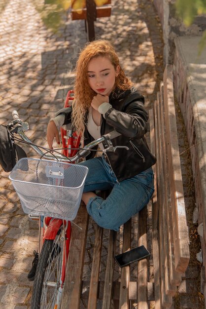 Joven rubia sentada en un banco, descansando en el parque con su bicicleta