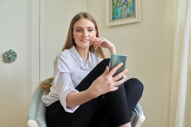 Joven rubia sentada en casa en una silla usando comunicación por video en un teléfono inteligente