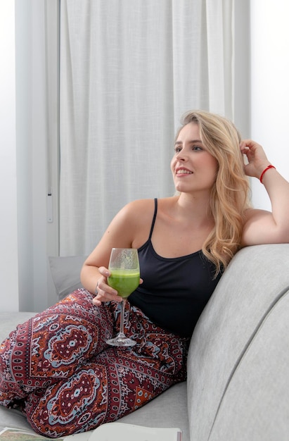 Joven rubia sentada en el sofá bebiendo un batido de verduras