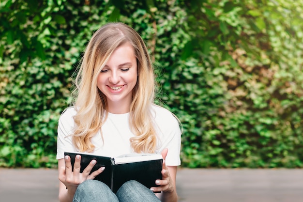 Joven rubia se sienta en un banco, lee un libro y sonríe en un parque