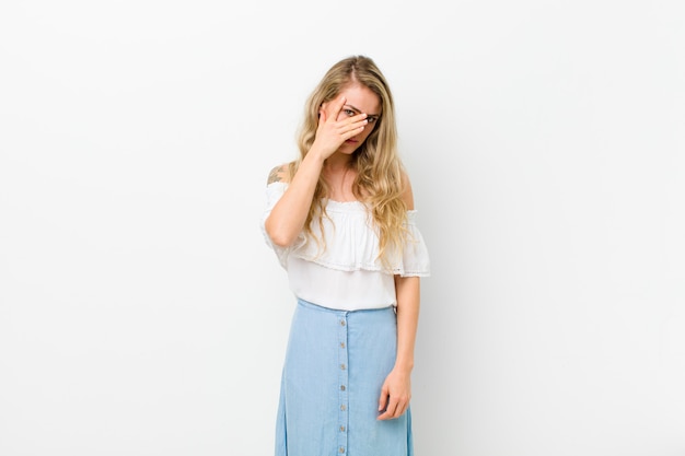 Joven rubia se siente aburrida, frustrada y con sueño después de una tarea aburrida, aburrida y tediosa, sosteniendo la cara con la mano en la pared blanca