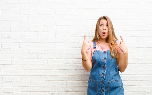 Joven rubia se siente asombrada y con la boca abierta apuntando hacia arriba con una mirada sorprendida y sorprendida sobre la pared de ladrillo