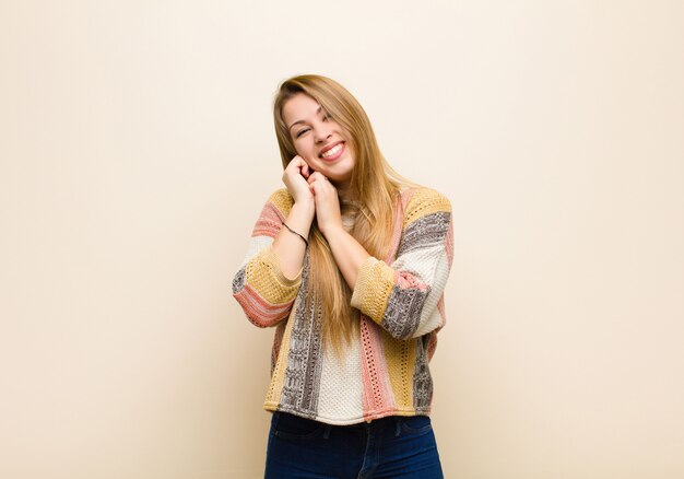 Joven rubia se siente enamorada y se ve linda, adorable y feliz, sonriendo románticamente con las manos junto a la cara contra la pared de color beige