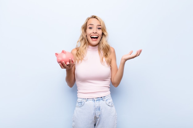 Joven rubia se siente feliz, emocionada, sorprendida o sorprendida, sonriendo y asombrada por algo increíble sosteniendo una alcancía