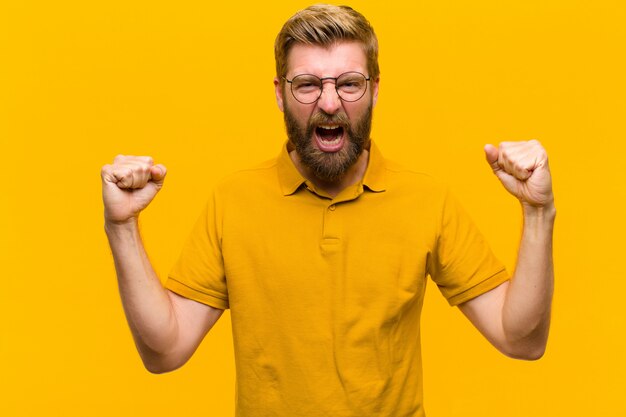 Joven rubia se siente feliz, positiva y exitosa, celebrando la victoria, los logros o la buena suerte en la pared naranja