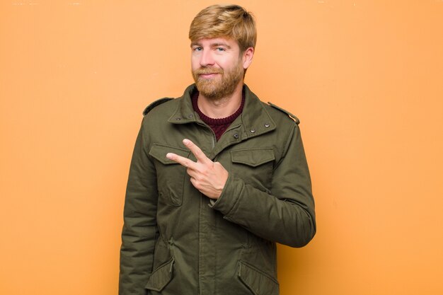 Joven rubia se siente feliz, positiva y exitosa, con la mano haciendo forma de V sobre el pecho, mostrando la victoria o la paz contra la pared plana