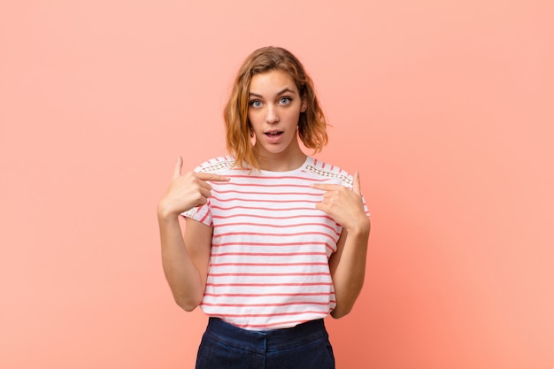 Joven rubia se siente feliz, sorprendida y orgullosa, apuntando a sí misma con una mirada emocionada y asombrada en la pared de color plano