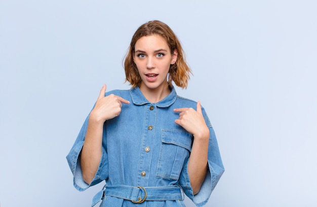 Joven rubia se siente feliz, sorprendida y orgullosa, apuntando a sí misma con una mirada emocionada y asombrada sobre la pared de color