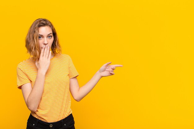 Joven rubia se siente feliz, sorprendida y sorprendida, cubriendo la boca con la mano y apuntando al espacio de copia lateral contra la pared de color plano