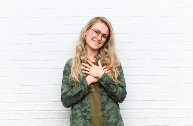 Joven rubia se siente romántica, feliz y enamorada, sonriendo alegremente y cogidos de la mano cerca del corazón contra el fondo de la pared de ladrillo