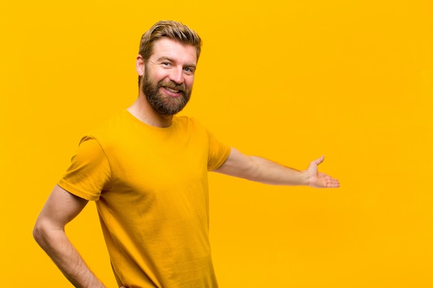 Joven rubia sintiéndose feliz y alegre, sonriendo y dándote la bienvenida, invitándote a entrar con un gesto amistoso contra la pared naranja