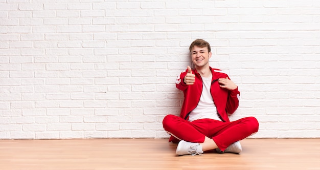 Joven rubia sintiéndose orgulloso, despreocupado, confiado y feliz, sonriendo positivamente con los pulgares arriba sentados en el suelo