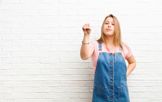 Joven rubia sintiéndose seria, fuerte y rebelde, levantando el puño, protestando o luchando por la revolución contra la pared de ladrillo