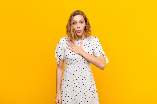 Joven rubia sintiéndose sorprendida y sorprendida, sonriendo, tomando la mano al corazón, feliz de ser la única o mostrando gratitud contra la pared de color plano